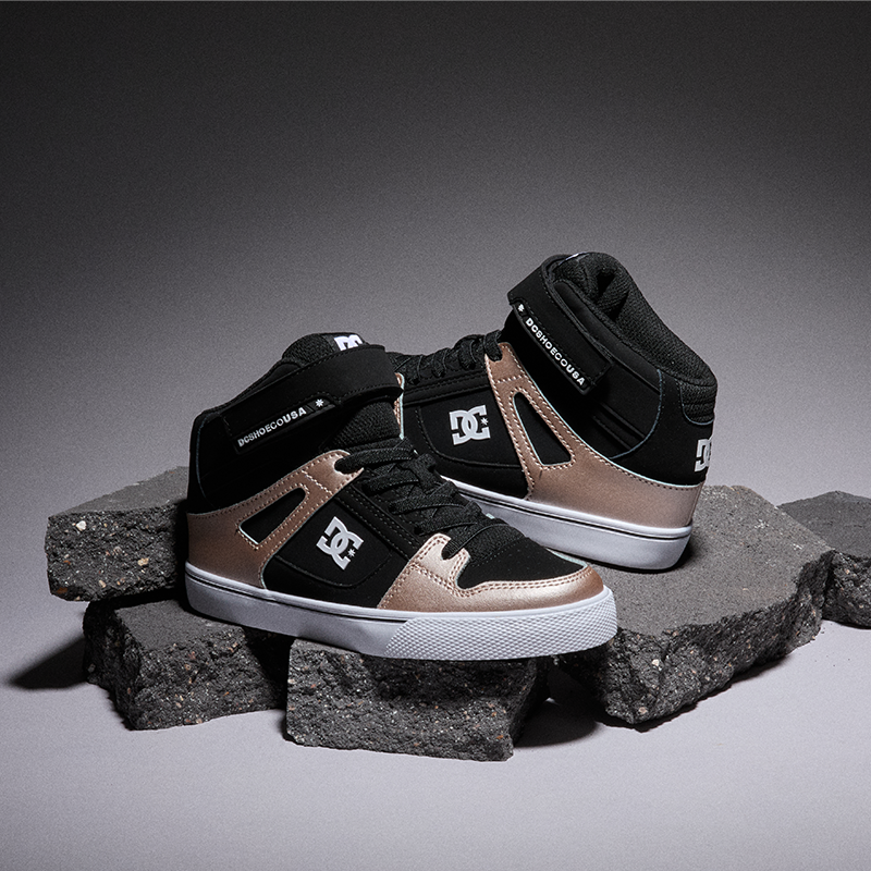 A pair of DC Shoes high-top sneakers in black and metallic bronze, displayed on dark gray stone blocks against a dark gray studio background.