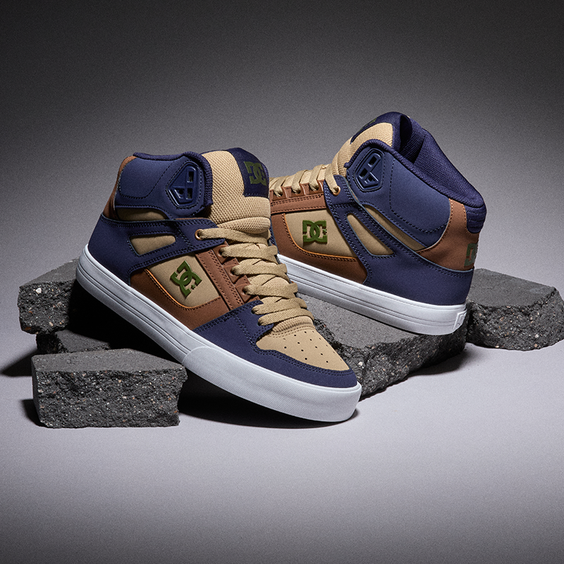 A pair of DC high-top sneakers in navy blue, tan, and brown, displayed on stacked dark gray concrete blocks against a dark gray studio background.