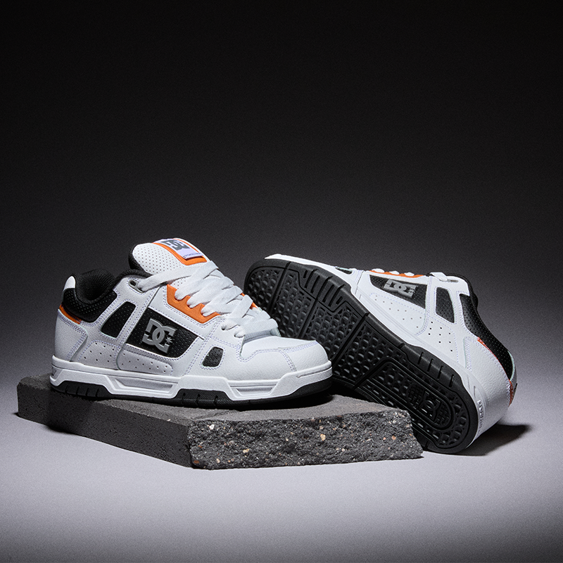 A pair of DC sneakers in white, black, and orange, with one shoe standing upright on a dark rectangular block and the other lying on its side showing the sole, set against a dark gray studio background.