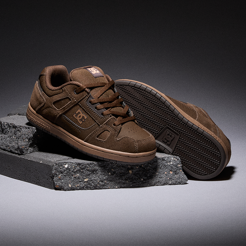 A pair of DC brand low-top sneakers in brown suede and synthetic materials, displayed on dark textured stone blocks against a dark gray studio background.