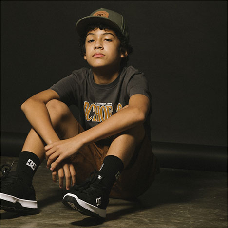 Boy wearing a cap, t-shirt, and shorts in white and black DC sneakers sitting on the ground against a dark background