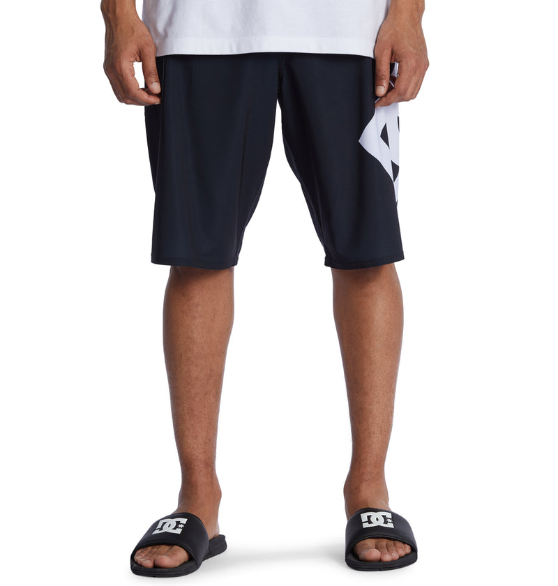 A person wears Reebok Lanai 21 black shorts with a large white graphic on the left leg, a white T-shirt, and black DC slide sandals. Only the lower half of their body is visible.