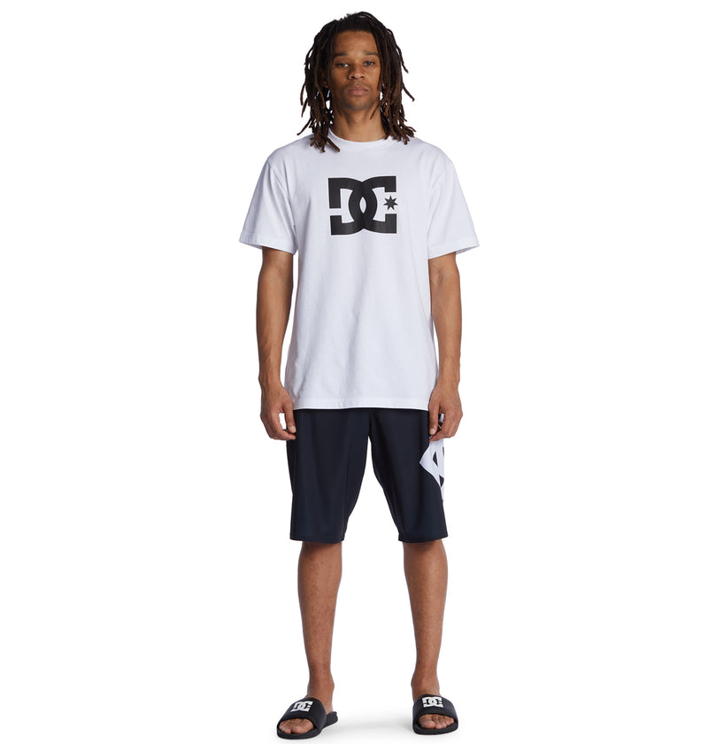 A man with long dreadlocks wears the white Reebok Lanai 21 t-shirt, black shorts with a white patch, and black slide sandals, standing against a plain white background.