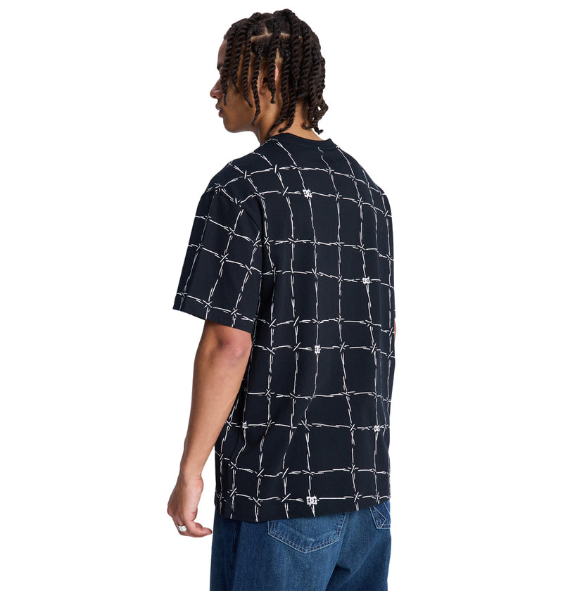 A person with braided hair, seen from the back, models the Reebok DC Wired Resort Hss—a loose black T-shirt with a white barbed wire grid pattern—paired with blue jeans against a plain white background.