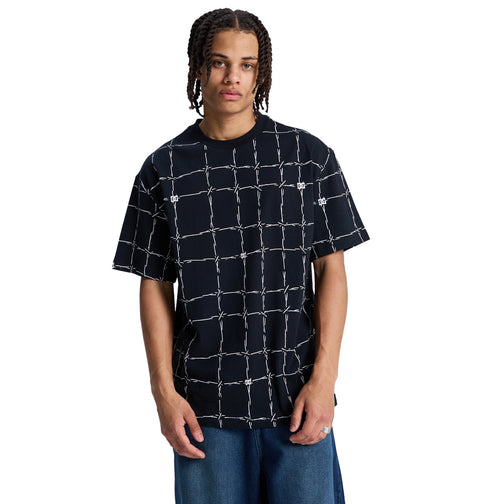 A person with braided hair wears the Reebok DC Wired Resort Hss, a loose black T-shirt with a white grid pattern, styled with blue jeans and posed against a plain white background.