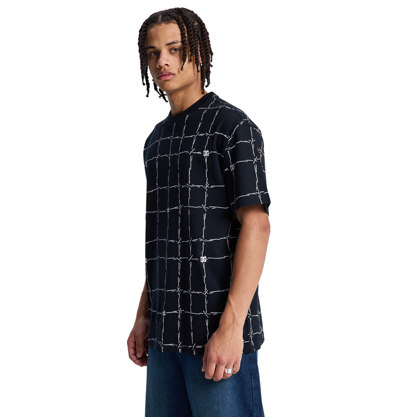 A young man with braided hair models the Reebok DC Wired Resort Hss black T-shirt featuring a white barbed wire grid pattern and blue jeans, standing sideways and looking at the camera against a white background.