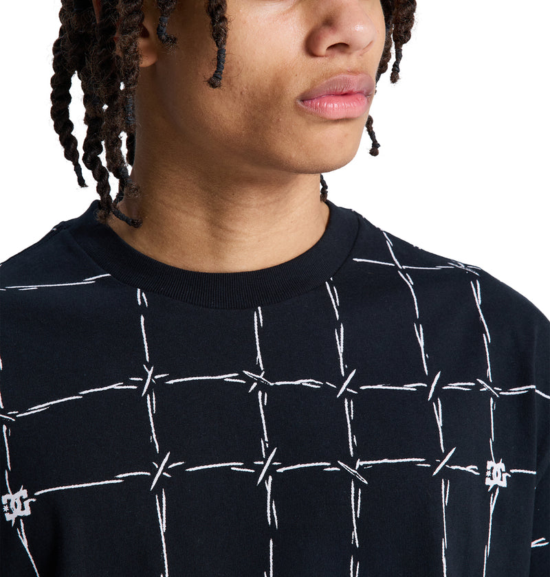 A person with medium brown skin and short, twisted hair wears a Reebok DC Wired Resort Hss black shirt featuring a white barbed wire pattern. The photo is from the shoulders up with their face partially visible, looking slightly to the side.