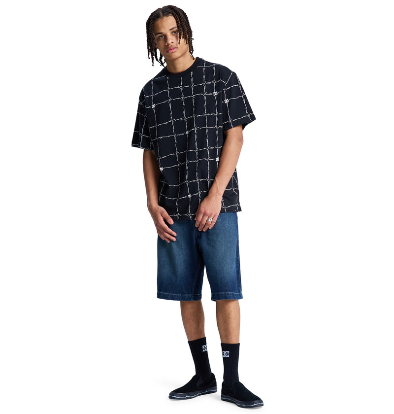 A person wears the Reebok DC Wired Resort Hss black t-shirt with a white grid pattern, paired with knee-length denim shorts, black socks featuring white text, and black slip-on shoes against a plain white background.