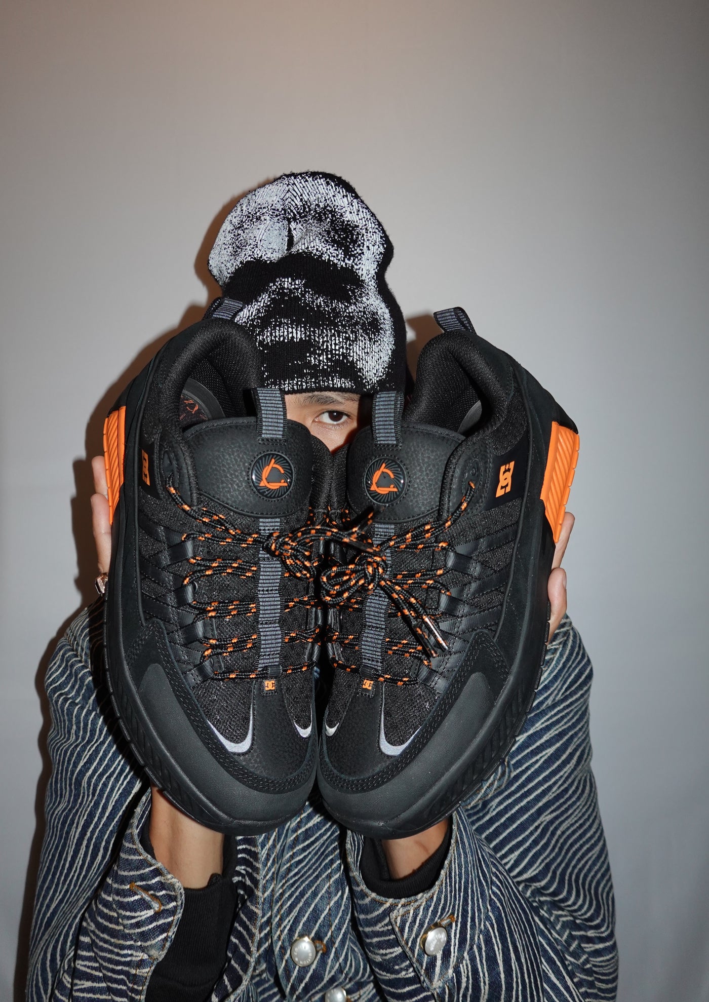 Person holding black sneakers with orange accents and a knit beanie against a plain background