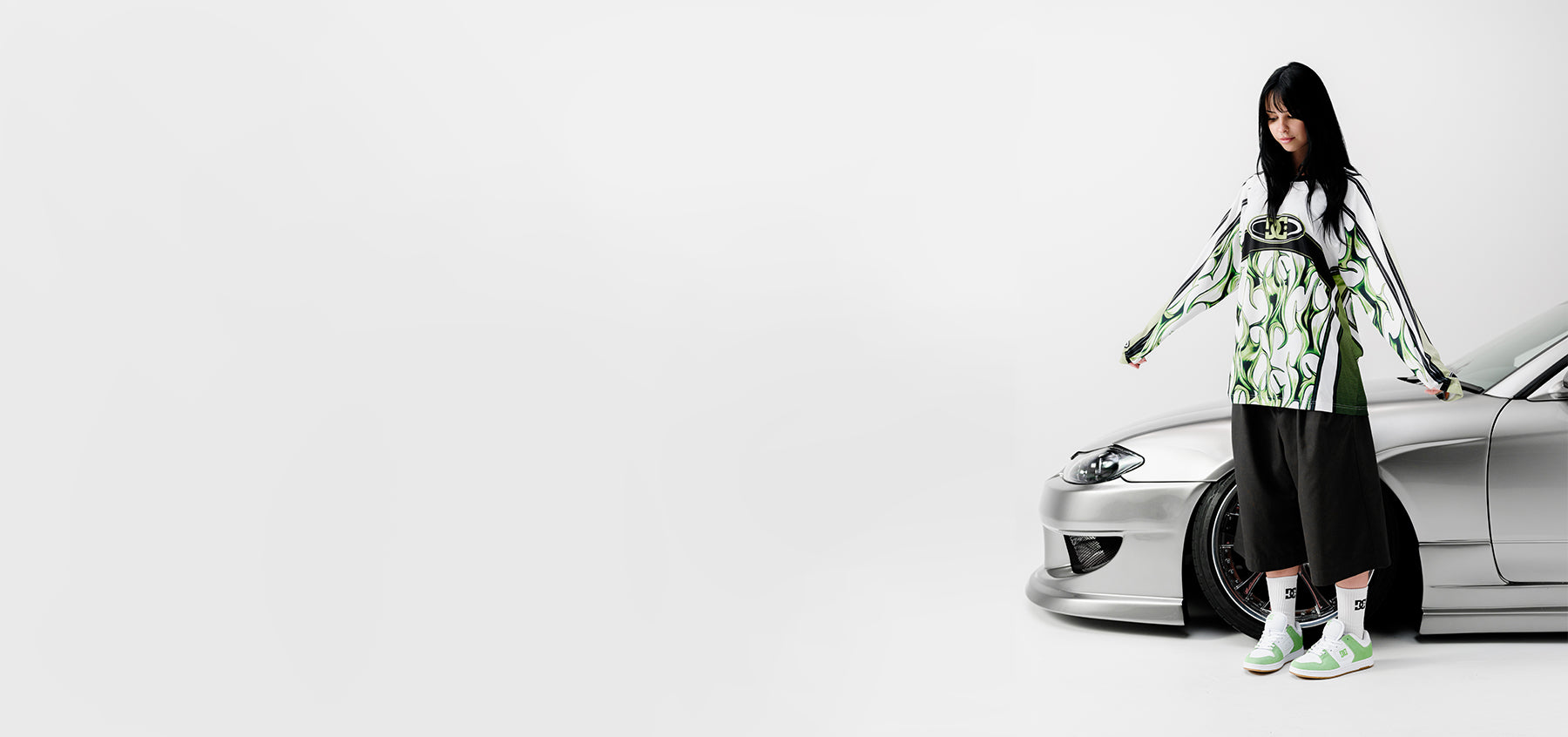 Person in white and green shirt, black pants, and green and white sneakers standing against silver car on a white background