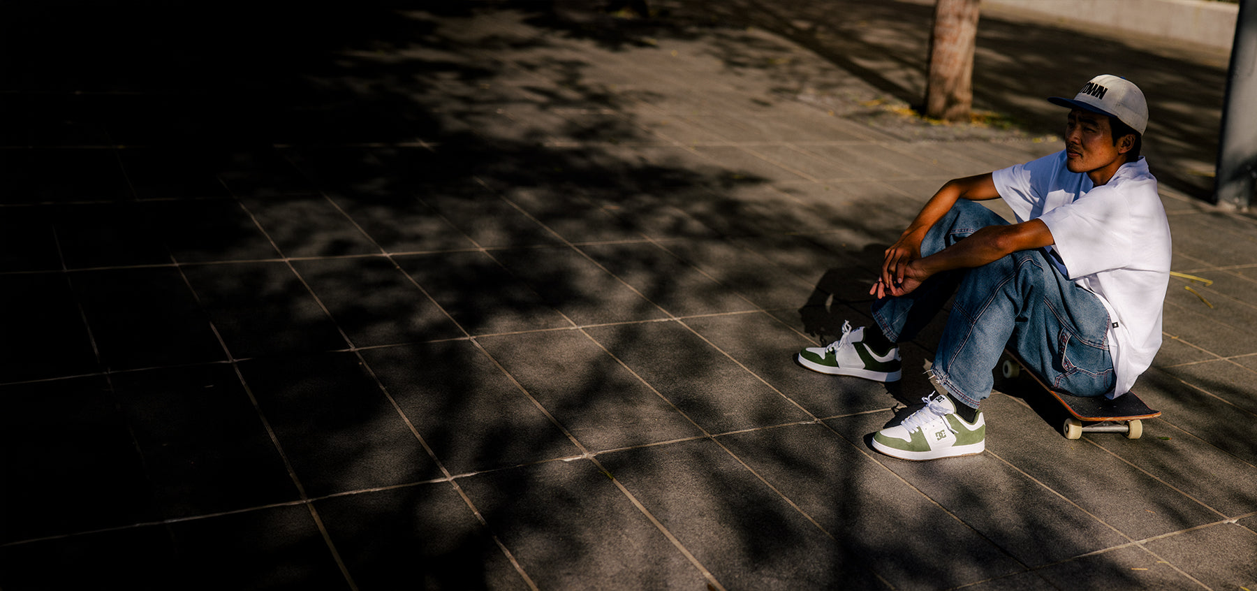 Person in a white t-shirt, jeans, and green and white sneakers sitting on skateboard outside