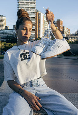 Person wearing a white t-shirt with a logo, holding white sneakers in an urban setting.