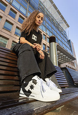 Person wearing white sneakers with black accents, sitting on a bench in an urban setting.