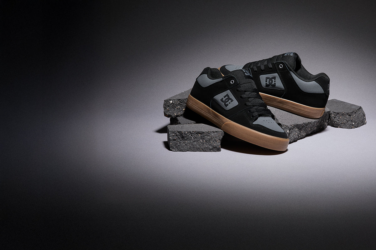 A pair of DC Shoes low-top sneakers in black and gray suede with gum soles, displayed on stacked dark concrete blocks against a dark gray studio background.