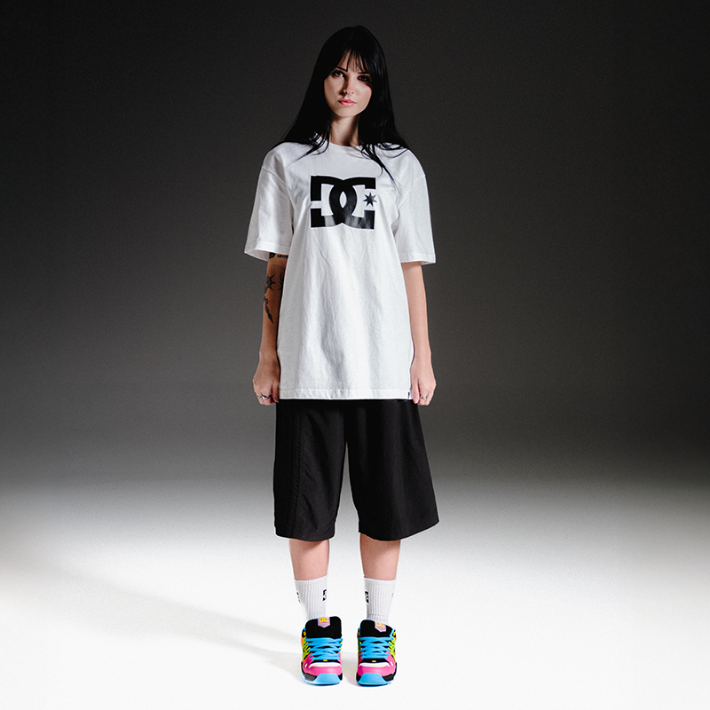 A model stands against a dark gray gradient studio background wearing an oversized white DC t-shirt with a black logo, black shorts, white socks, and colorful sneakers.