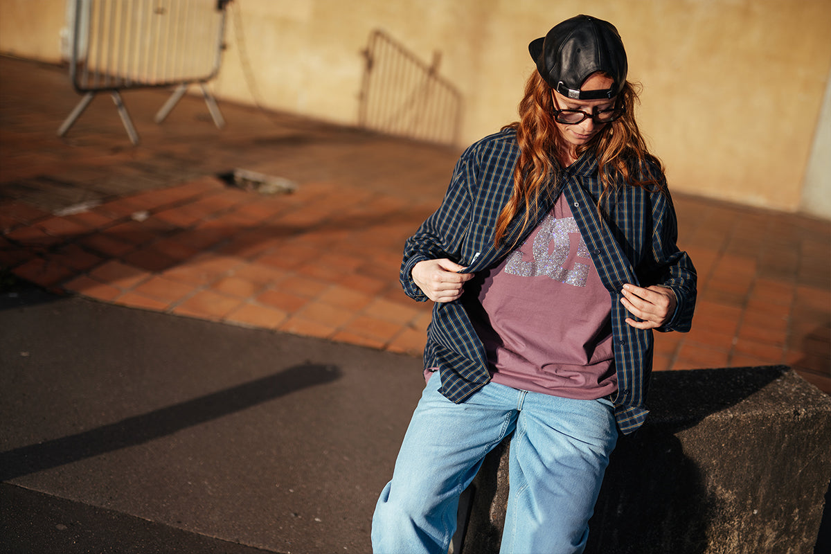 A person wearing a black baseball cap, sunglasses, a dark blue plaid flannel overshirt over a mauve graphic t-shirt, and light blue jeans, sitting on a concrete block in an outdoor urban setting.
