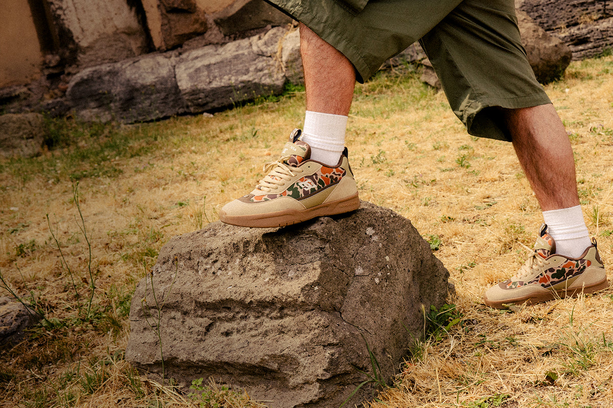 A person wearing beige and multicolor patterned sneakers with white socks, standing outdoors on a large rock in a grassy, stone-filled environment.