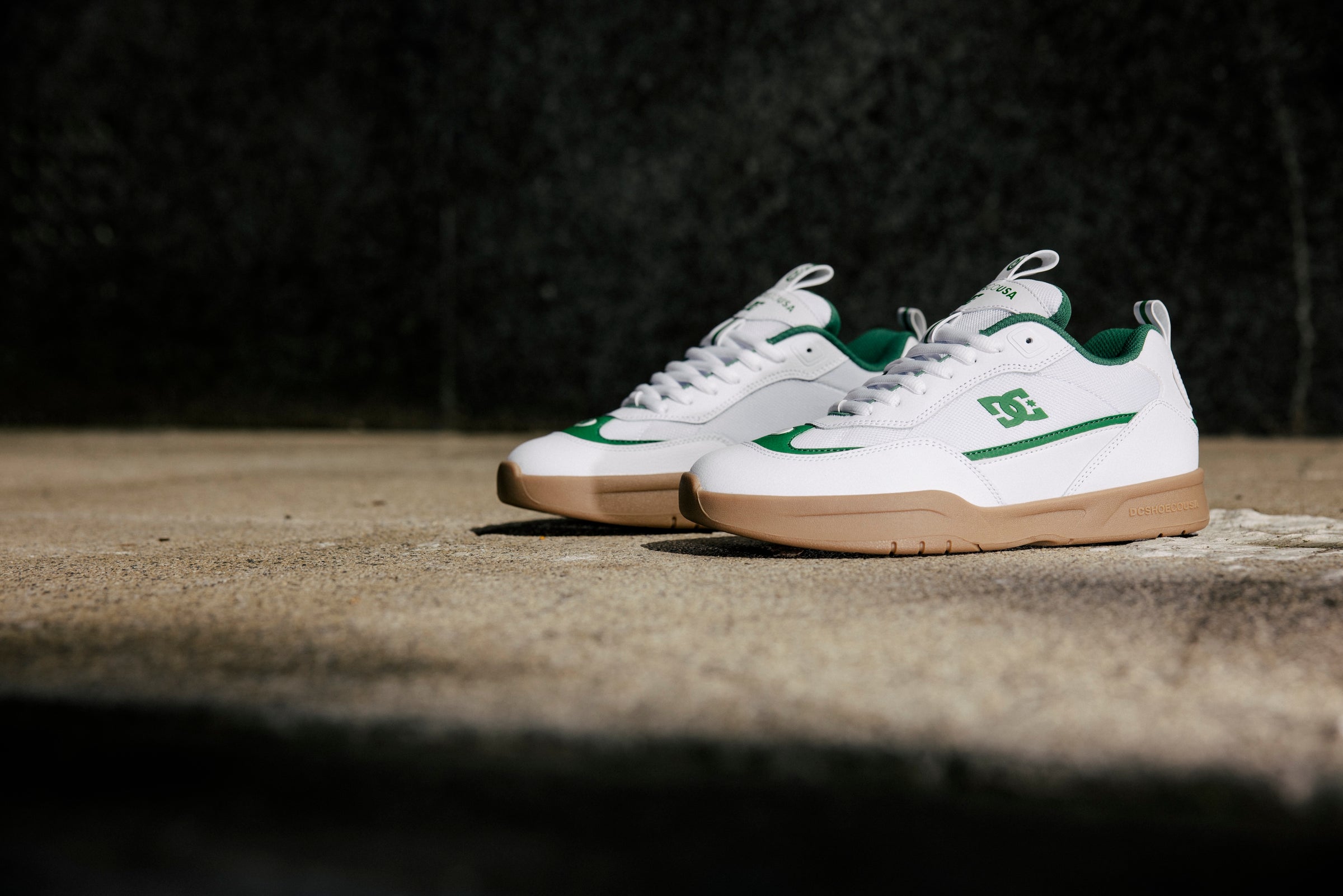 A pair of white and green DC brand sneakers with gum soles placed side by side on a sunlit concrete surface, with a dark background.