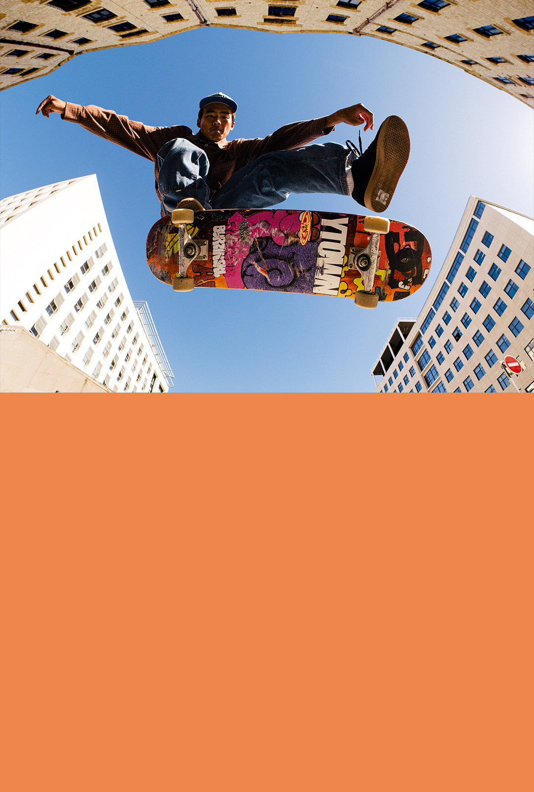 Skateboarder performing a trick in an urban setting with buildings in the background above orange colorblock