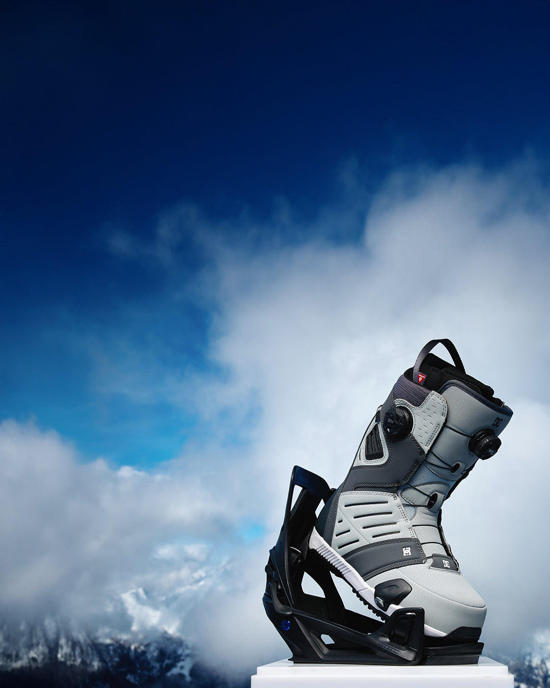 Gray and black snowboard boot with bindings against a snowy mountain background