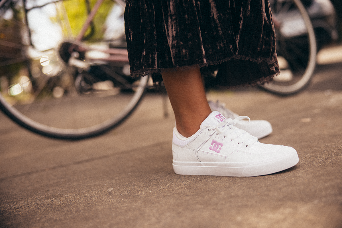 White sneakers with a pink logo worn by a person on a street with a blurred bicycle in the background.