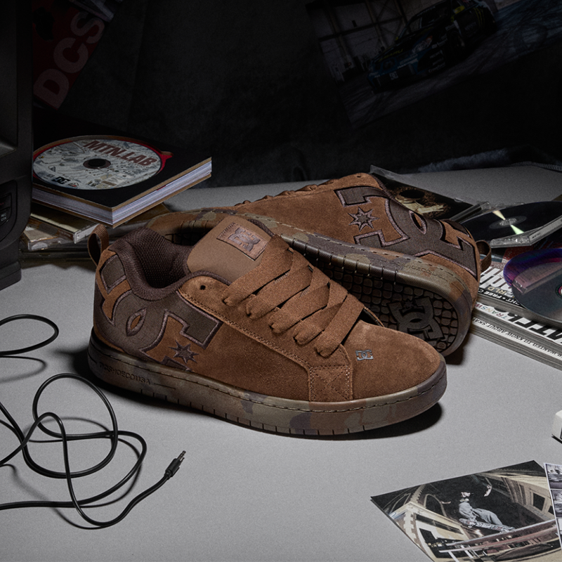 A pair of low-top brown suede skate shoes with distressed detailing and star motifs, displayed on a gray tabletop surrounded by various dark-themed props including a CD, cables, and printed materials.