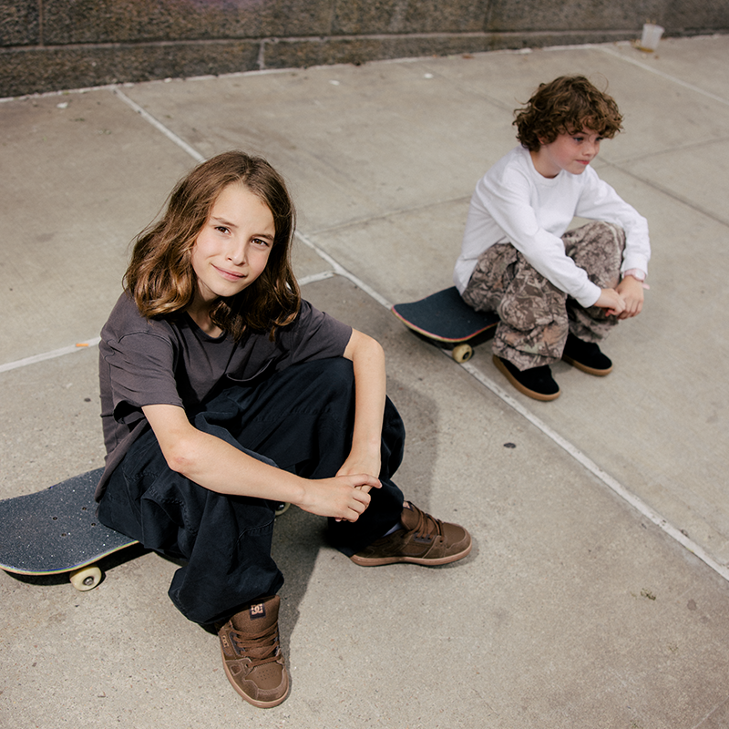 Two young skateboarders sitting on concrete outdoors. The child in the foreground wears a dark gray t-shirt, dark pants, and brown sneakers, sitting on a skateboard.