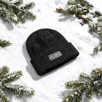 Black knit beanie with DC logo on a snowy surface with pine branches.