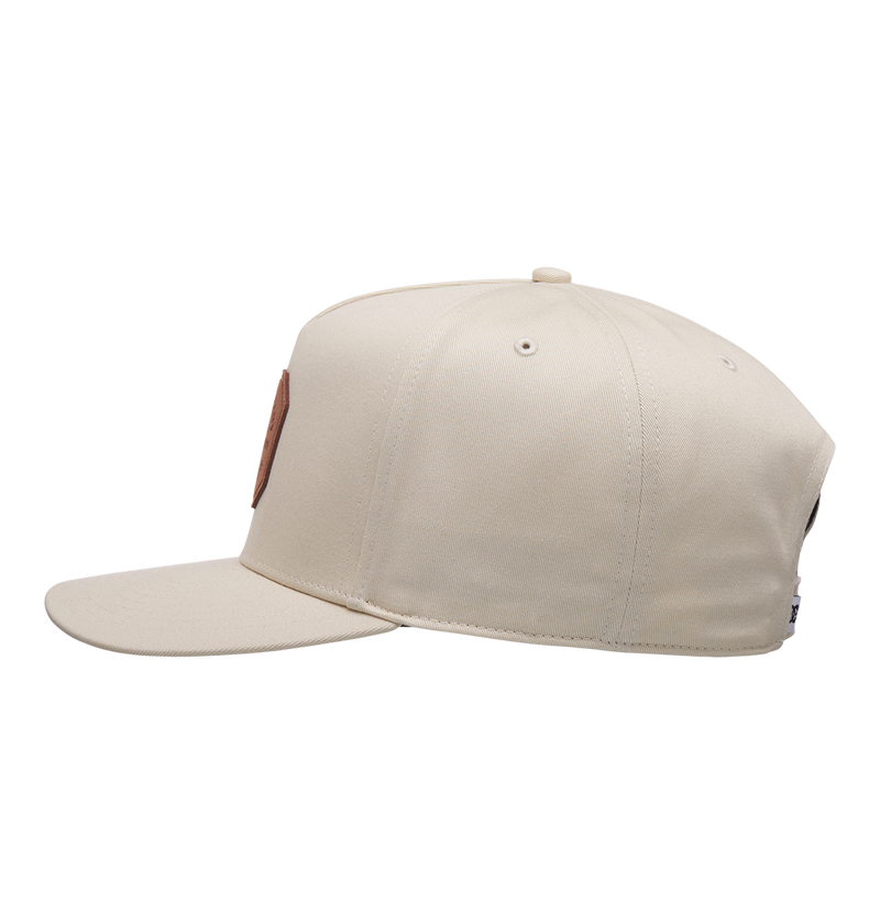 The Men's Reynotts Cap by DC Shoes is displayed side-on against a white background. This beige skate cap features a round brown DC patch on the front and visible stitching along its panels.