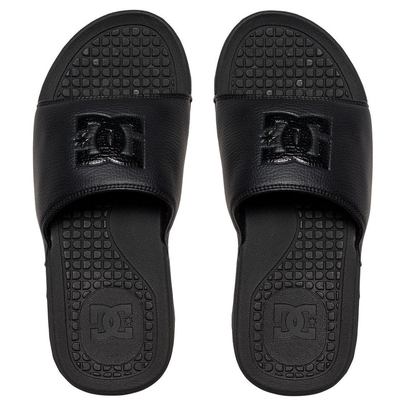 A pair of DC Shoes Men's Bolsa Slides in black, featuring wide straps with a raised DC logo and textured soles, are placed side by side and viewed from above.