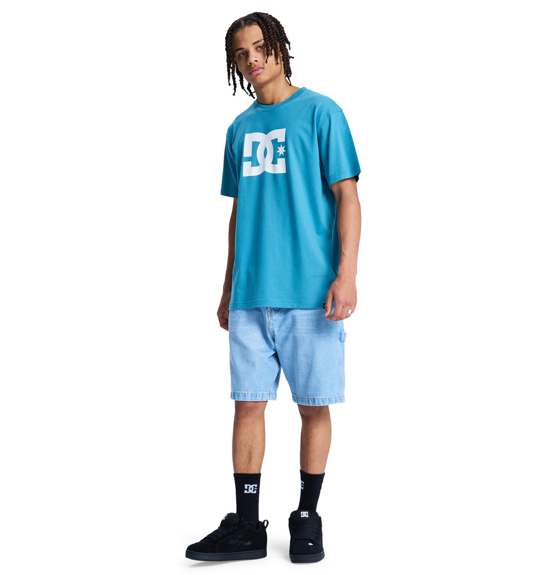 A person with braided hair, wearing a blue Reebok DC Star Hss t-shirt, light denim shorts, black socks, and black sneakers stands facing forward against a plain white background.