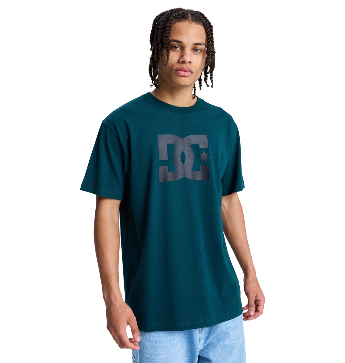 A person with medium-length braided hair wears a teal Reebok DC Star Hss t-shirt and light blue jeans, standing against a plain white background.