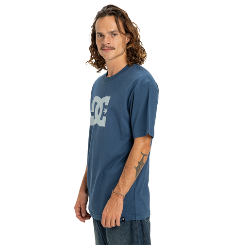 A man with long wavy hair and a mustache wears the Men's DC Star Tee by DC Shoes, showing his right forearm tattoo and gazing at the camera with a neutral expression against a white background.