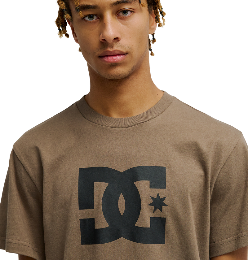 A young person with light brown skin and blonde-tipped dreadlocks wears the men's DC Star Tee by DC Shoes, a relaxed fit brown skate shirt with a bold black DC Shoes graphic on the chest, standing against a plain white background.