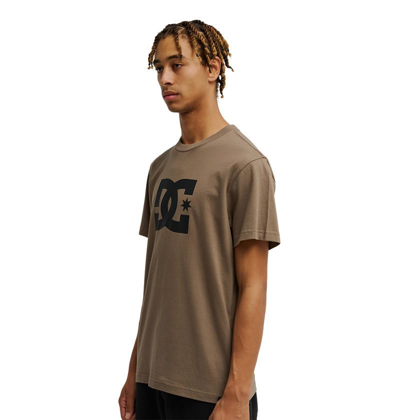 A person with short, blond-tipped braids wears a relaxed fit brown Men's DC Star Tee by DC Shoes, featuring a large black logo on the chest, standing against a plain white background.