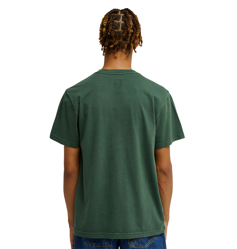 A person with short, twisted hair is shown from the back against a plain white background, wearing the DC Shoes Men's DC Star Pigment Dye Tee in dark green paired with blue jeans.