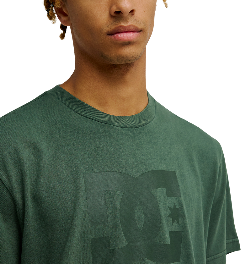 A person is shown from the shoulders up against a plain background, wearing the Men's DC Star Pigment Dye Tee by DC Shoes—a green skate t-shirt featuring a subtle logo on the front and a relaxed fit.