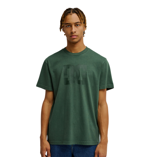 A person with short, curly hair is wearing the Men's DC Star Pigment Dye Tee by DC Shoes—a dark green graphic skate shirt featuring a subtle DC artwork design and a relaxed fit—while standing against a plain white background.