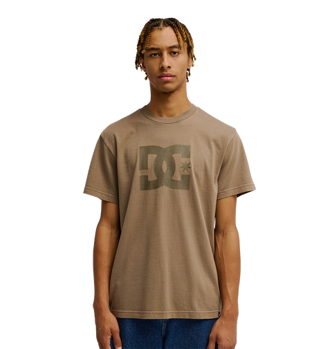 A young man with short braids is wearing the Men's DC Star Pigment Dye Tee from DC Shoes, styled with blue jeans and standing against a plain white background.