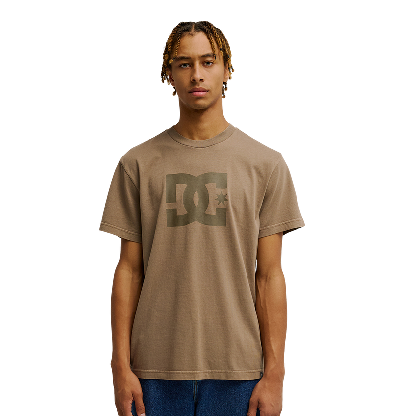 A young man with short braids is wearing the Men's DC Star Pigment Dye Tee from DC Shoes, styled with blue jeans and standing against a plain white background.