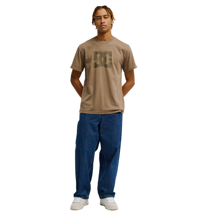 A person with short, blonde braids wears the Men's DC Star Pigment Dye Tee from DC Shoes—a relaxed fit brown skate t-shirt featuring iconic DC branding—paired with loose blue jeans and white sneakers, standing with hands behind their back against a plain white background.