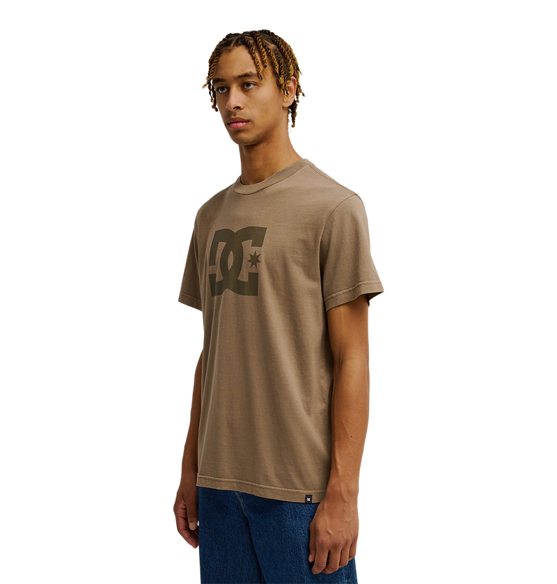 A young man with short, twisted hair wears the Men's DC Star Pigment Dye Tee by DC Shoes—a taupe graphic skate shirt featuring signature DC artwork—paired with blue jeans. He stands against a plain white background, gazing slightly to the side with a neutral expression.
