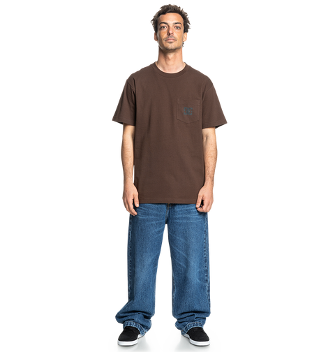 A man faces forward in a relaxed fit brown Men's DC Star Pocket Tee by DC Shoes, featuring a chest pocket, paired with loose blue jeans and black skate shoes against a plain white background.