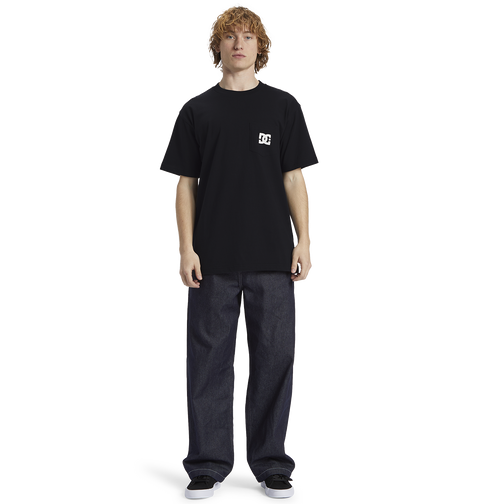 A person with curly hair wears the Men's DC Star Pocket Tee by DC Shoes in black, paired with relaxed fit dark jeans and black shoes, standing against a plain white background.