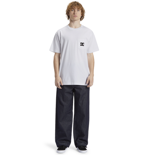 A person with curly hair wears the Men's DC Star Pocket Tee by DC Shoes—a relaxed fit white cotton skate shirt with a small logo—paired with wide dark jeans and black shoes, standing before a plain white background.