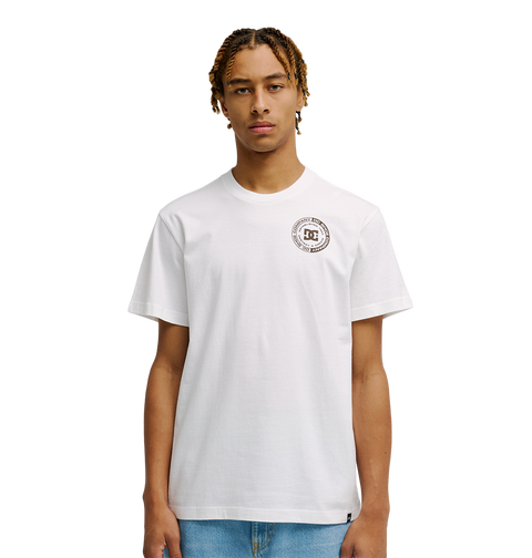 A young man with short, braided hair models the Men's DC Corpo Tee by DC Shoes—a relaxed fit, plain white t-shirt with a small black DC logo and circular emblem on the left chest—paired with light blue jeans against a white background.