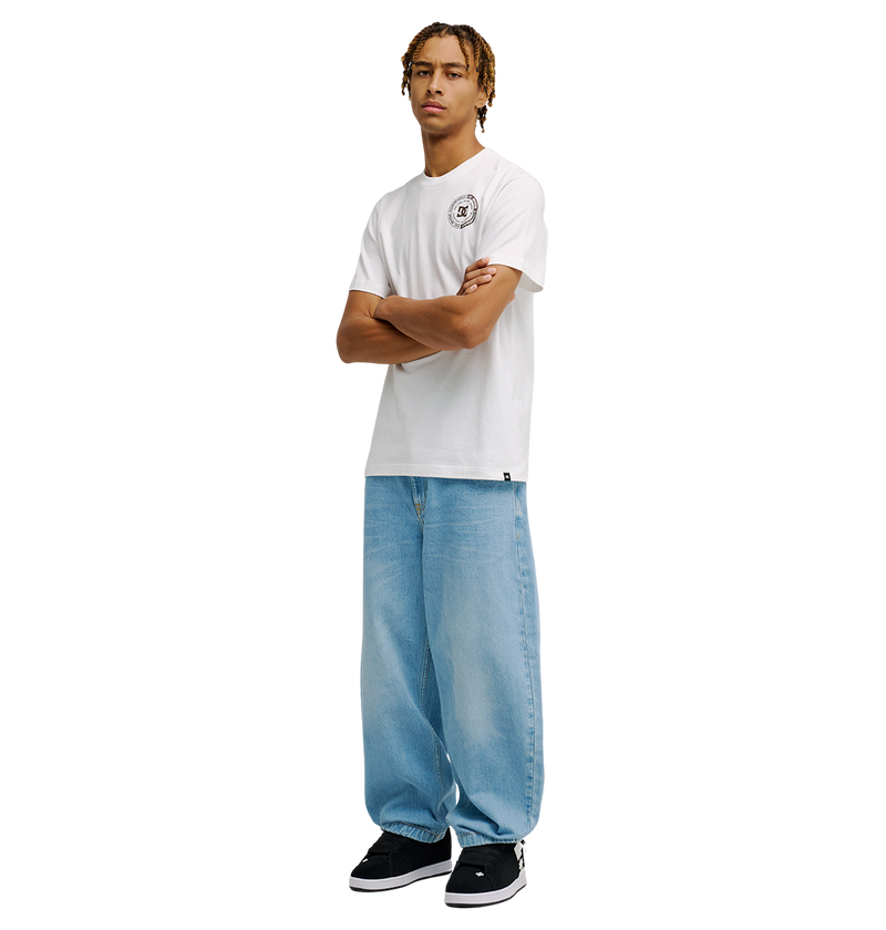 A person stands with arms crossed, wearing the Men's DC Corpo Tee by DC Shoes with a small circular logo on the chest, paired with light blue baggy jeans and black sneakers against a plain white background.