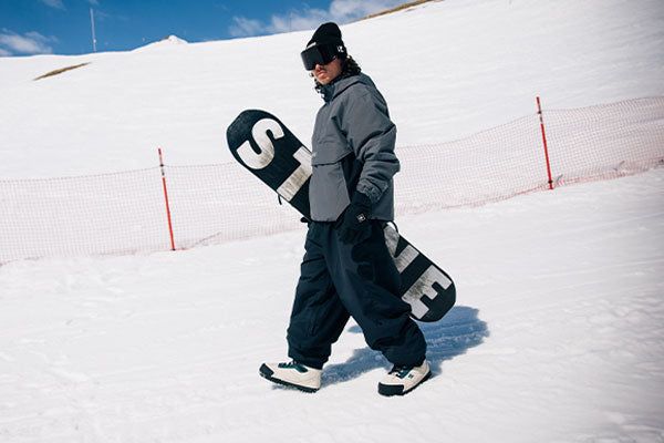 Person walking on a snowy slope with a snowboard, wearing a gray jacket and black pants.
