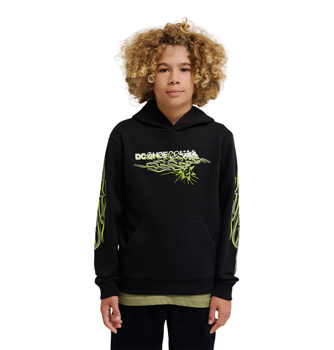 A young person with curly hair wears the DC Shoes Side Flame Hoodie Youth, a black pullover featuring neon green designs and a bold DC Shoes logo, standing against a plain white background.