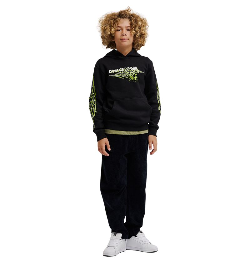 A child with curly hair wears the DC Shoes Side Flame Hoodie Youth, a black fleece hoodie with neon green DC graphics, paired with dark pants and white sneakers, standing facing forward against a plain white background.